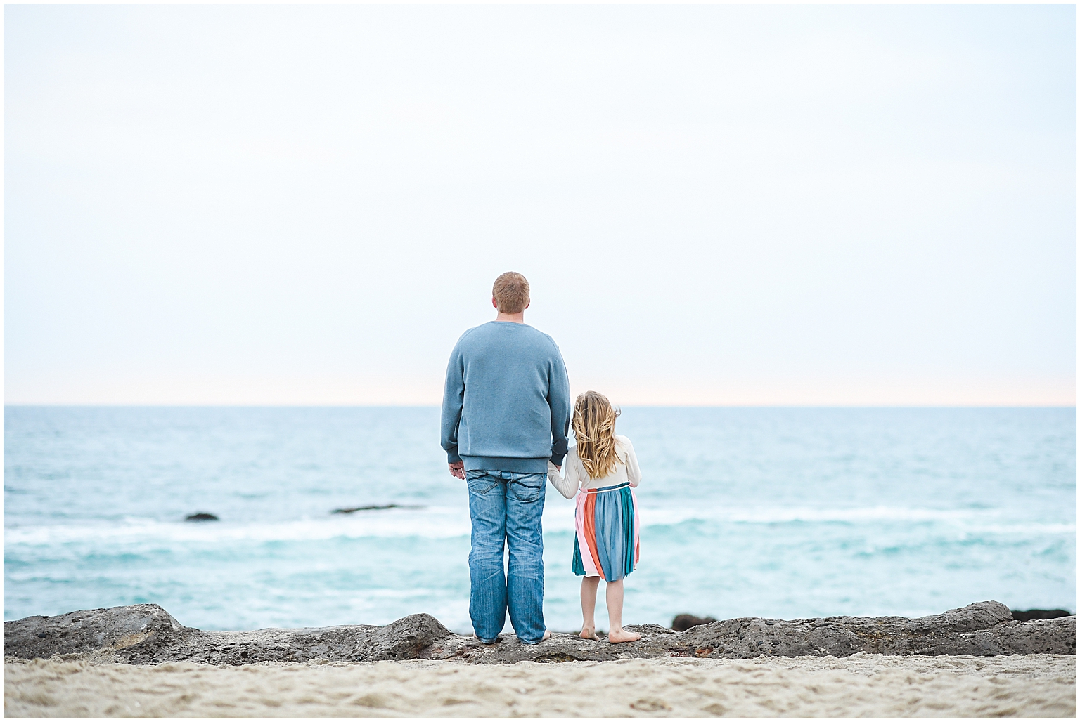 father and daughter look out at ocean while holding hands