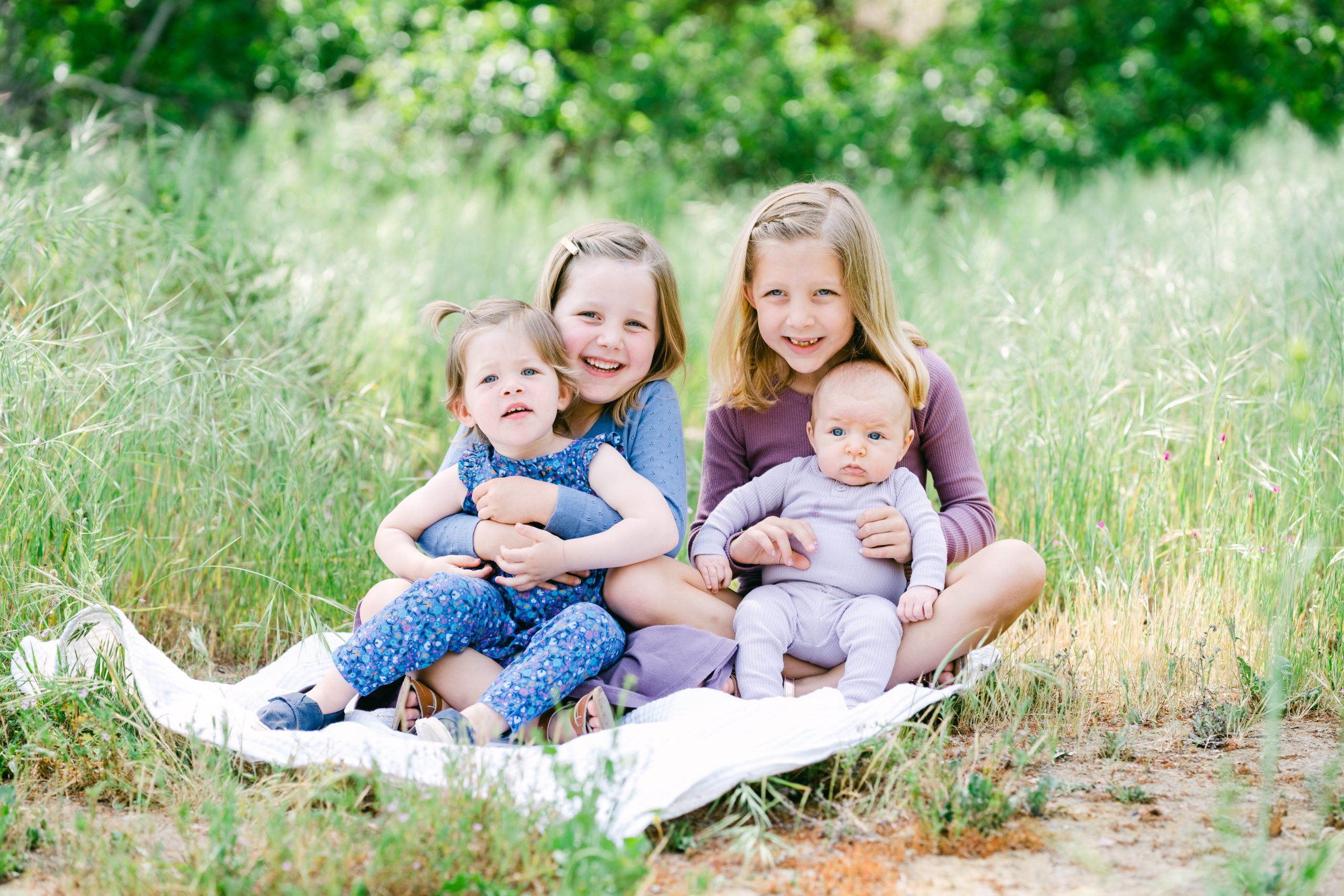 Four white children pose in the grassy fields of Marian Bear park. Three girls and a baby boy. The two older girls hold the younger one and the baby on their laps.