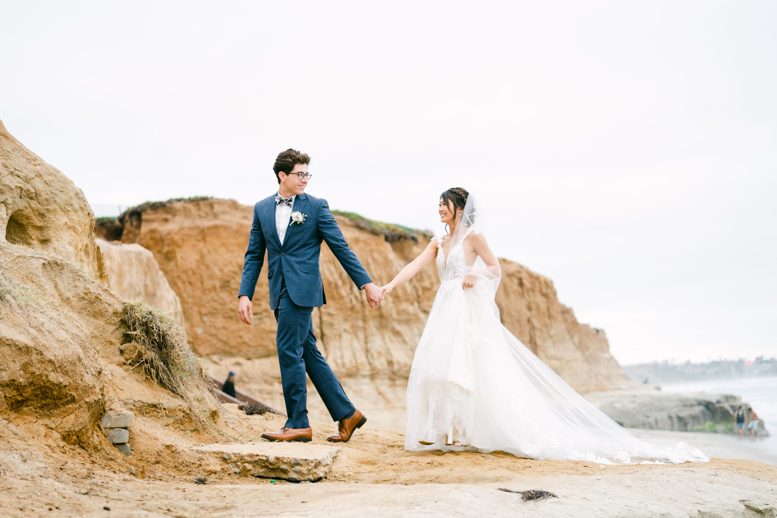 Wedding picture of a bride and groom, Aaron and Kara, on a Carlsbad, California beach. Dressed in a white wedding dress and a navy tux, they are holding hands and smiling at each other as they walk next to the cliffs. The photo was taken by Kate Noelle Photography.