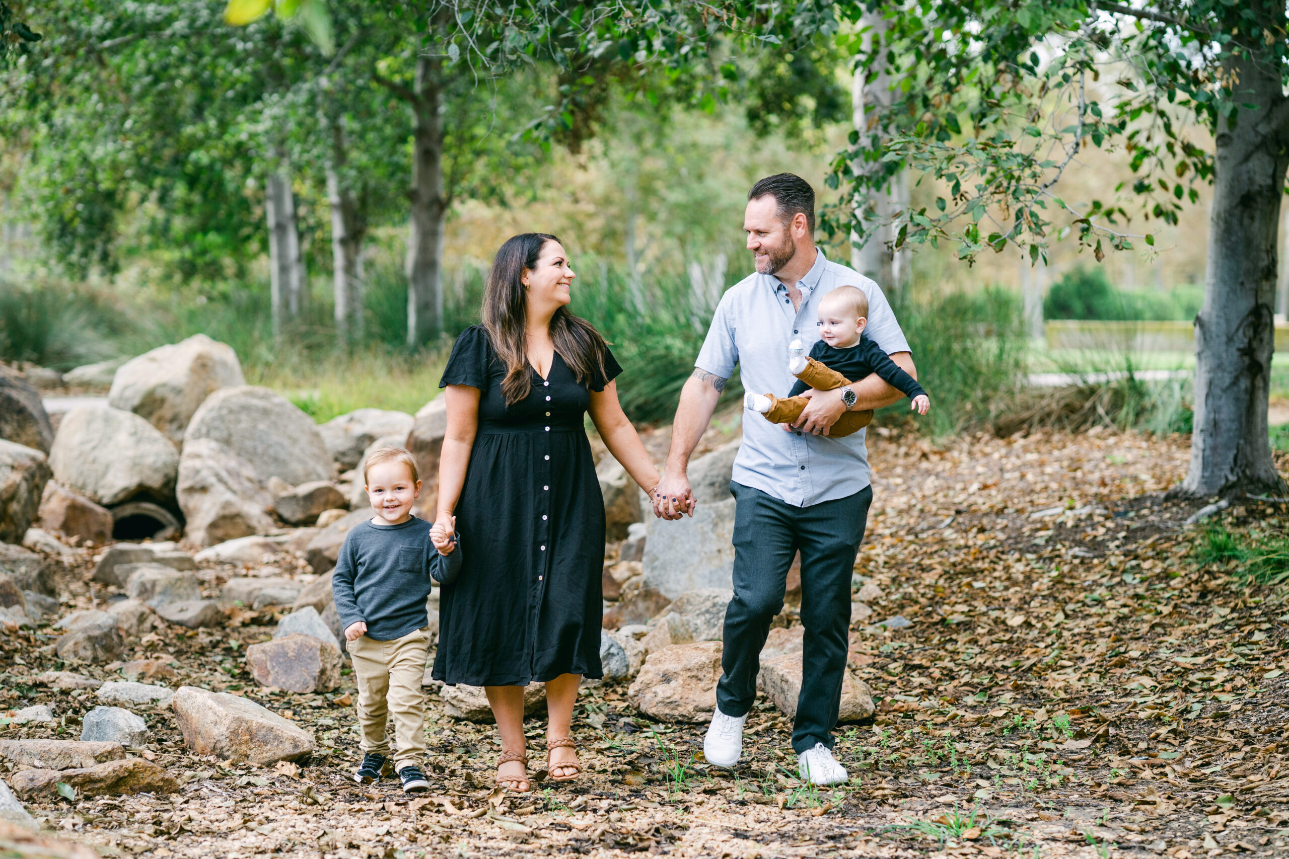 A white family of 4 poses on the Jeffrey Open Space Trail for family portraits in Irvine, California by Kate Noelle Photography. There is a mother, father, and their two boys-a toddler and a baby. They are all smiling at each other.