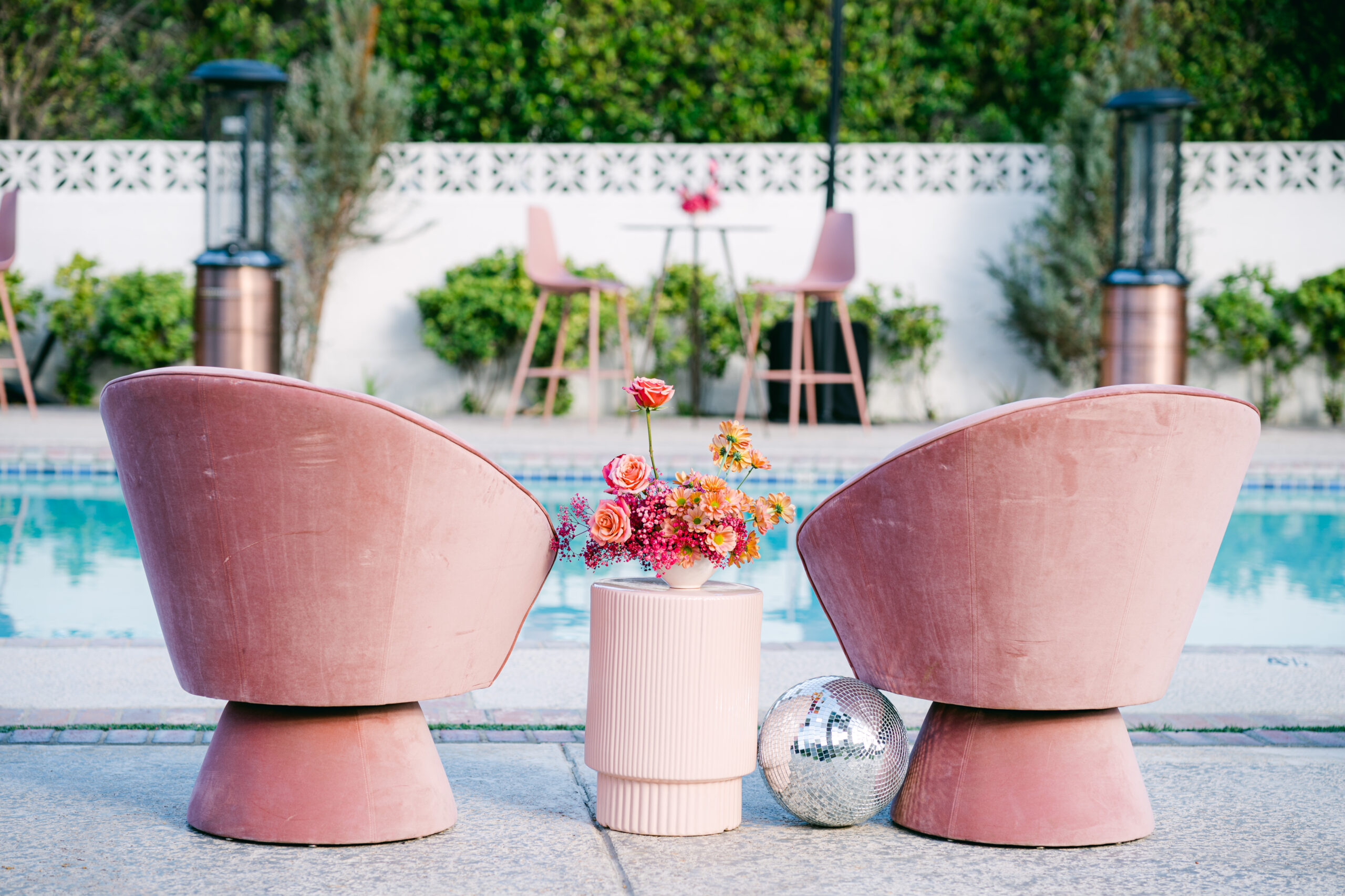 Cocktail hour setup by the pool for the Wedding Planner's Retreat hosted by California Wedding Day at the Ingleside Inn in Palm Springs. Two pink retro chairs on either side of a pink coffee table and floral arrangement, with a disco ball on the ground, designed by Nicole Alexandria Designs and photographed by Kate Noelle Photography