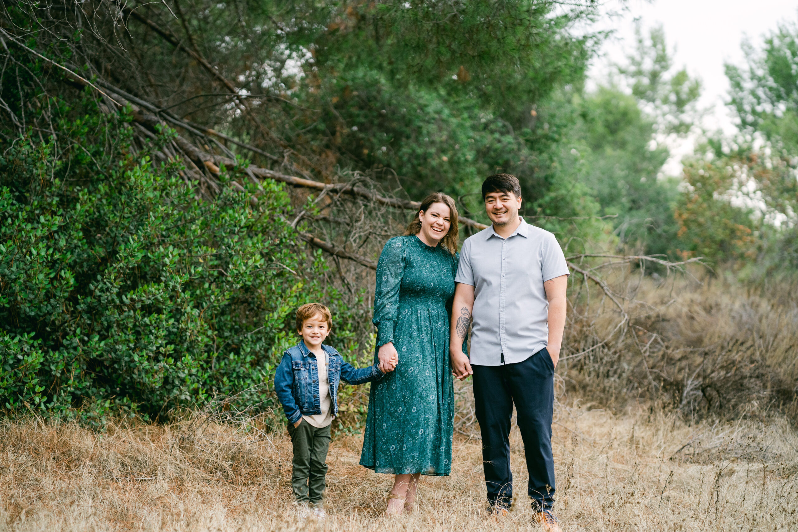 A family of three stands in Trabuco Canyon in Orange County. They are all holding hands and posing for family photos with Kate Noelle Photography.