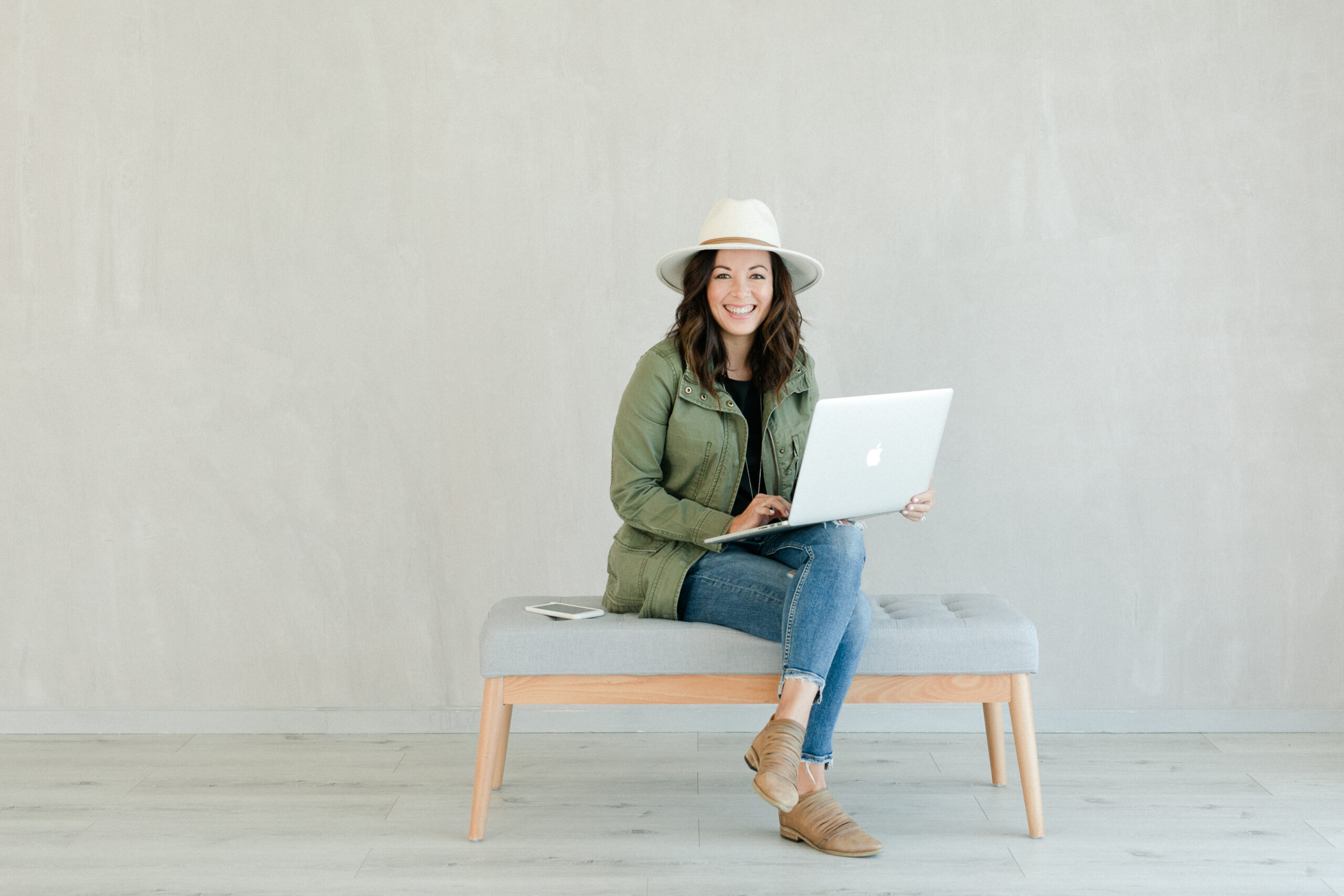 Best Amazon Prime Day photography deals of 2023 by Kate Noelle Photography: Kate sits on a bench with her white laptop and smiles. She is wearing jeans, a navy jacket, and a white wide-brimmed hat.