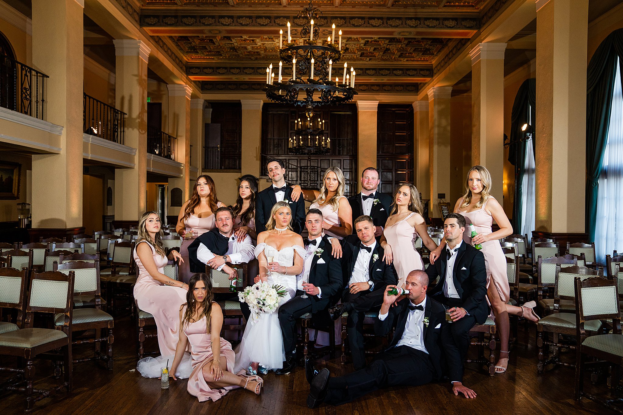 A large wedding party pose in a group at the the Wilshire Ebell of Los Angeles. They are posed like the promo photos from "Gossip Girl" and holding cocktails. The women are in light pink dresses and the men are in classic black suits. Photos by Kate Noelle Photography